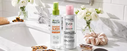 A clean and bright bathroom countertop/sink area featuring various hair accessories like a hair brush, hair clips, comb, scrunchie, etc. Also showing products mentioned in the article like: EverPure Glossing Treatment and Diamond Sleek Spray