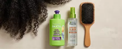 Frizzy hair pictured with Garnier Fructis Curl Nourish Curl Air Dry Cream and/or Diamond Sleek Shine-Coat Smoothing Spray and a hairbrush against a neutral background.