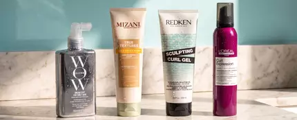 of Color Wow Anti-Frizz Spray for Curly Hair, Mizani Curl Enhancing Lotion, Redken Sculpting Curl Gel, and/or L'Oreal Paris Curl Expression 10-In-1 Cream-In-Mousse) lined up on a bathroom counter. A window featuring a blazing sun pictured as well.