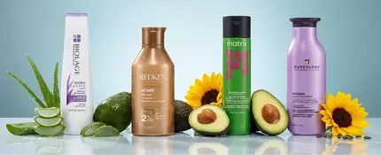 A lineup of four hydrating shampoos—Biolage, Redken, Matrix, and Pureology—arranged on a glossy surface surrounded by ingredients like aloe, avocado, and sunflower, visually highlighting moisture and softness.